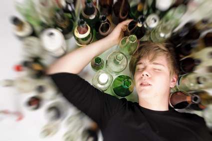 Junger Mann mit kurzen blonden Haaren liegt auf dem Boden und ist von vielen leeren Bier- und Schnapfsflaschen umgeben, Oberperspektive.